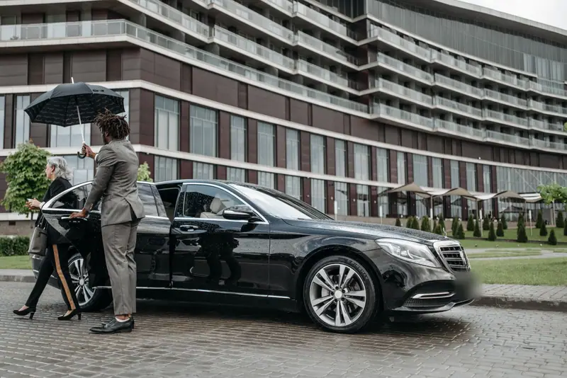 A chauffeur holds an umbrella over a passenger stepping out of a black Mercedes-Benz S-Class